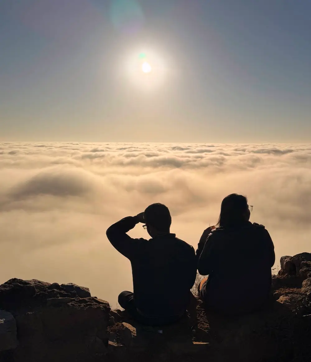 Two silhouettes sitting on a mountain summit above a sea of clouds